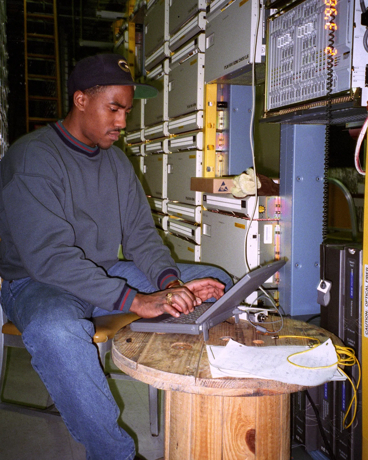 Engineer at a laptop on a cable spool in front of a wall of carrier transport equipment